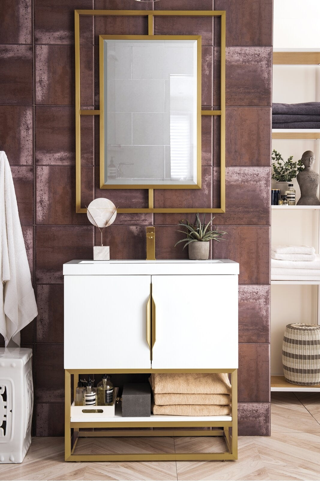 Freestanding modern white single-sink bathroom vanity with a stone countertop, gold faucet and hardware, open lower shelf with folded towels and bottles, and a matching gold-framed mirror against textured brown tile wall.