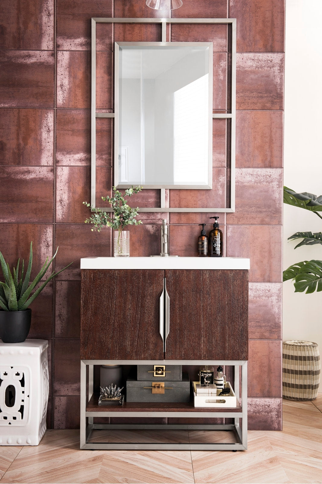Freestanding modern solid-wood bathroom vanity with a white stone top and single faucet, dark-stained cabinet doors on a metal base, open lower shelf with storage boxes, framed mirror and plants against rust-toned tile wall.