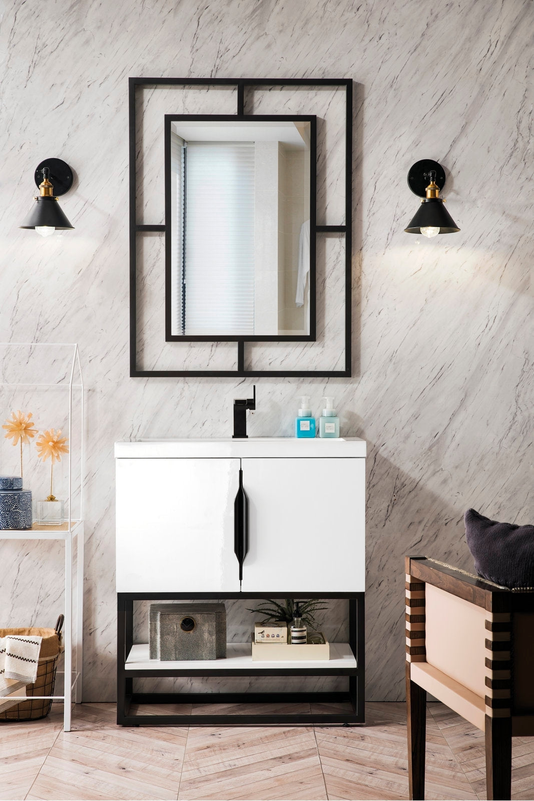 Modern white single-sink solid-wood vanity with a stone top, black single-handle faucet and vertical black handle, open black base shelf with decor, geometric black-framed mirror and two black-and-brass wall sconces on a marble-look wall.