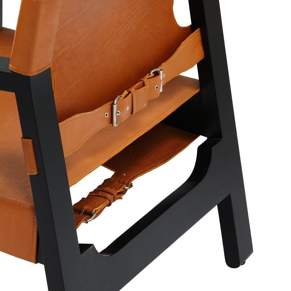 Close-up of tan leather seat and back straps with metal buckles attached to a matte black solid wood frame of a modern lounge chair.