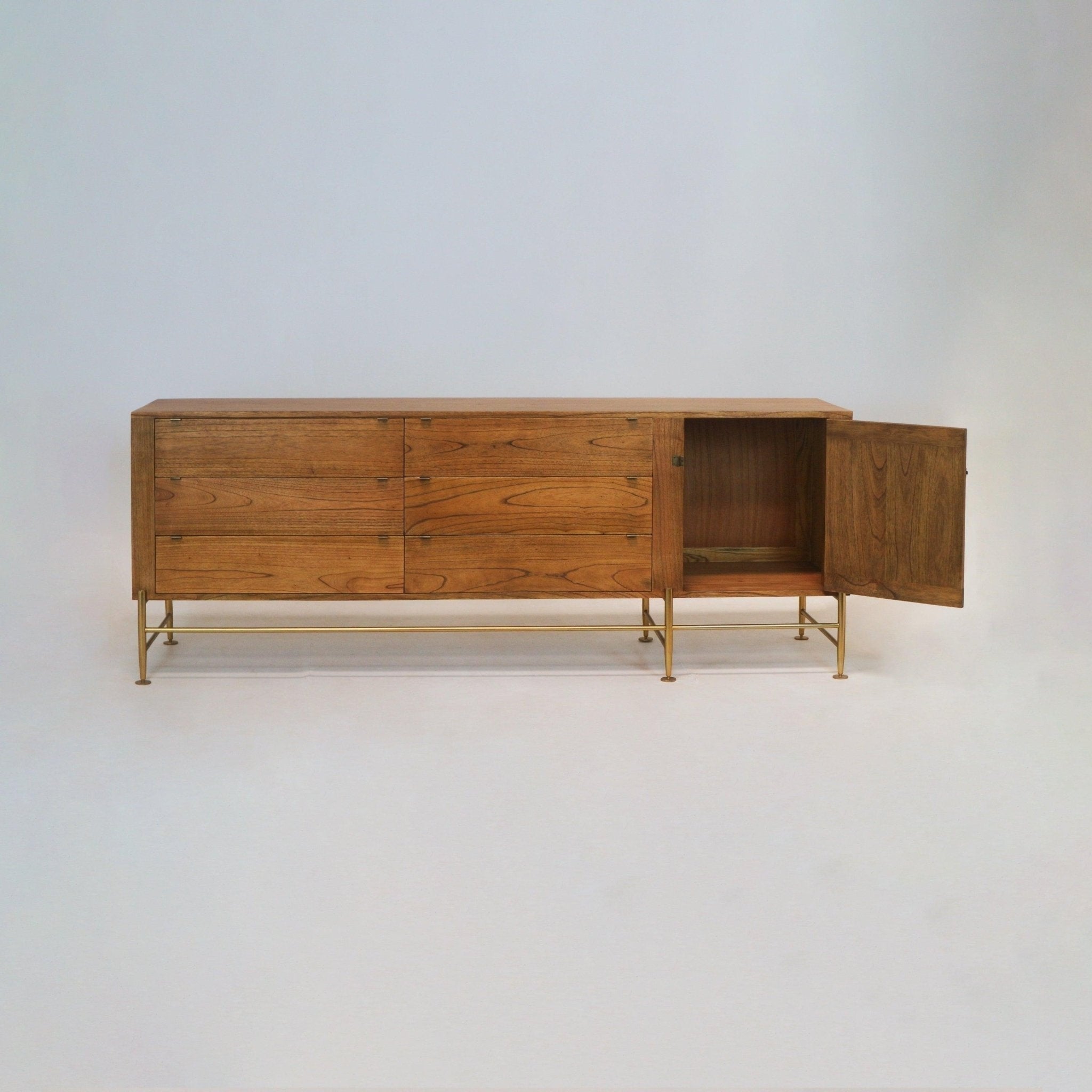 Long teak sideboard in mid-century modern style with six horizontal drawers and an open right cabinet door, raised on slim brass legs and crossbars, showing warm natural wood grain finish.