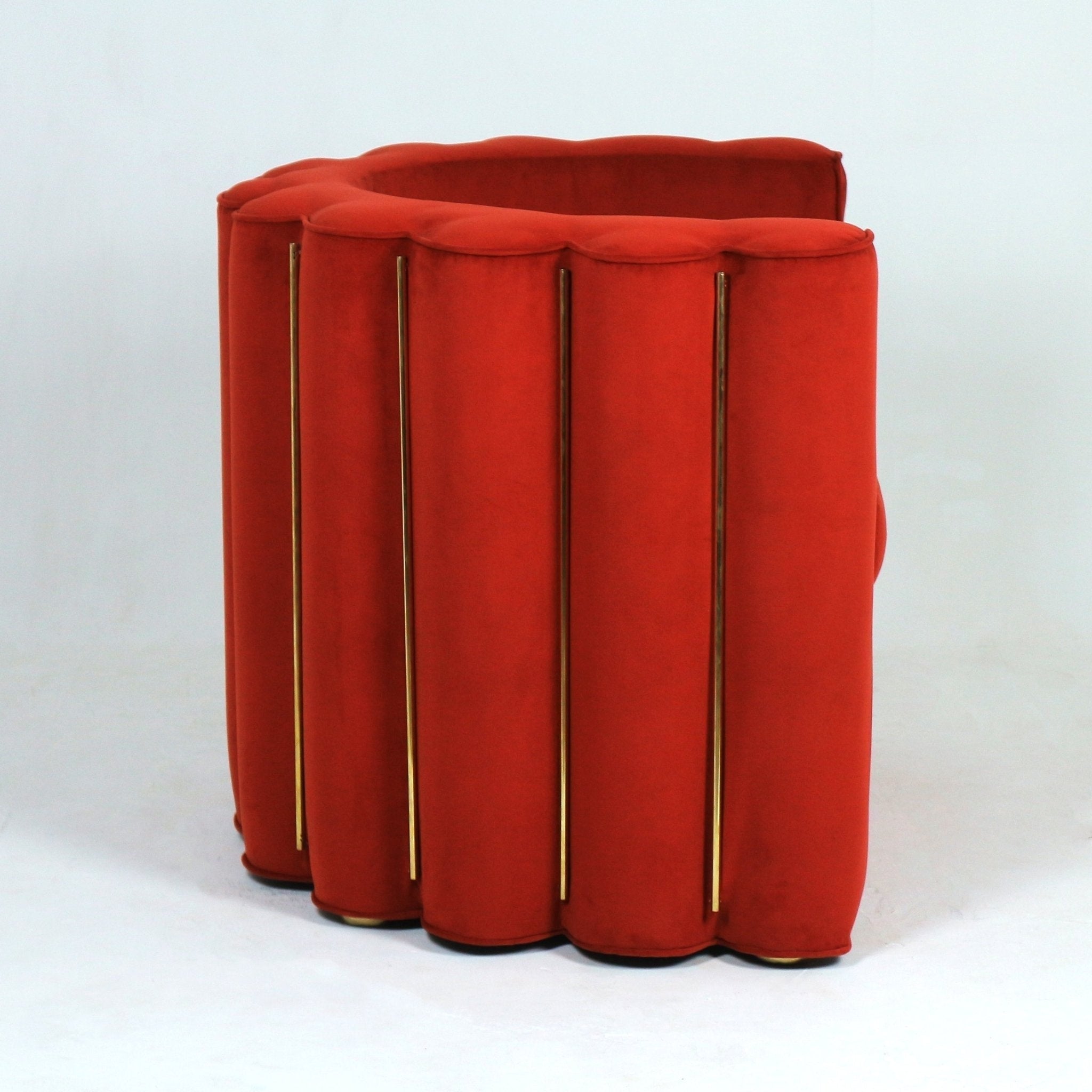 Three-quarter rear view of a modern red velvet fabric accent chair with a scalloped curved full back, plush upholstery and vertical brass rods along the exterior, resting on a low wooden base for living room seating.