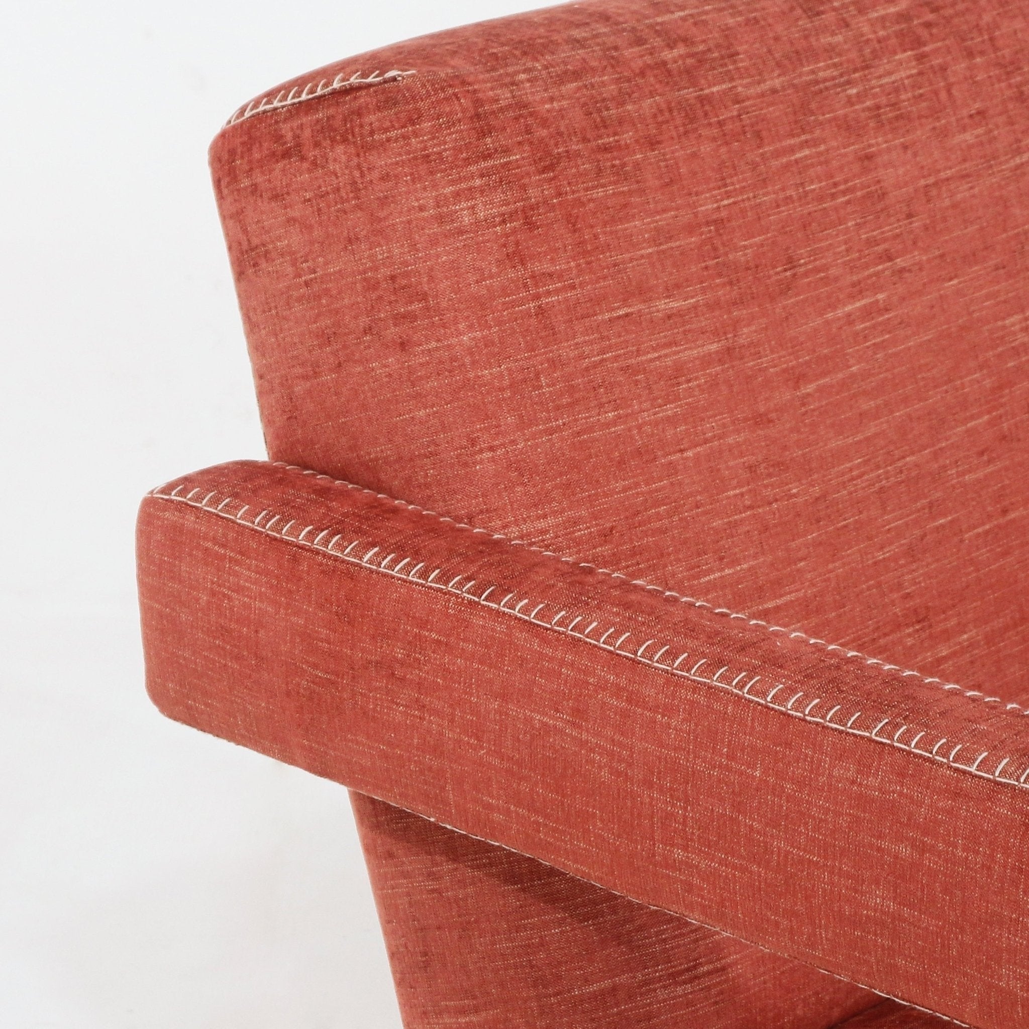 Close-up of a rust-colored fabric-upholstered modern lounge chair arm and back showing white contrast whipstitch detailing along the arm and top edge.
