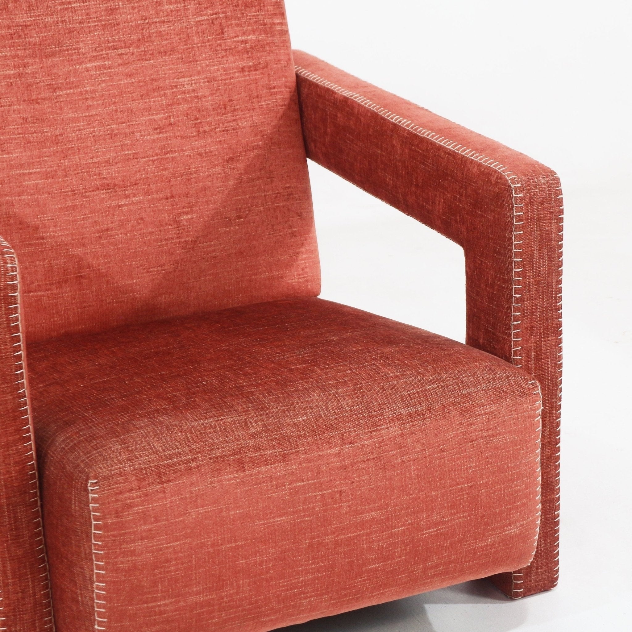 Close-up of a cabernet rust fabric-upholstered modern lounge chair showing a boxy arm and deep seat with white contrast stitching along the arm and seat edges and a wooden internal frame.