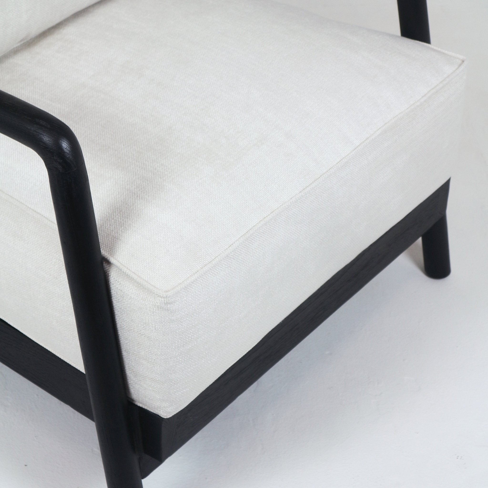 Close-up of a mid-century modern armchair seat showing a cream fabric cushion with piped edge resting on a dark-stained solid teak frame and rounded arm with exposed wooden base.