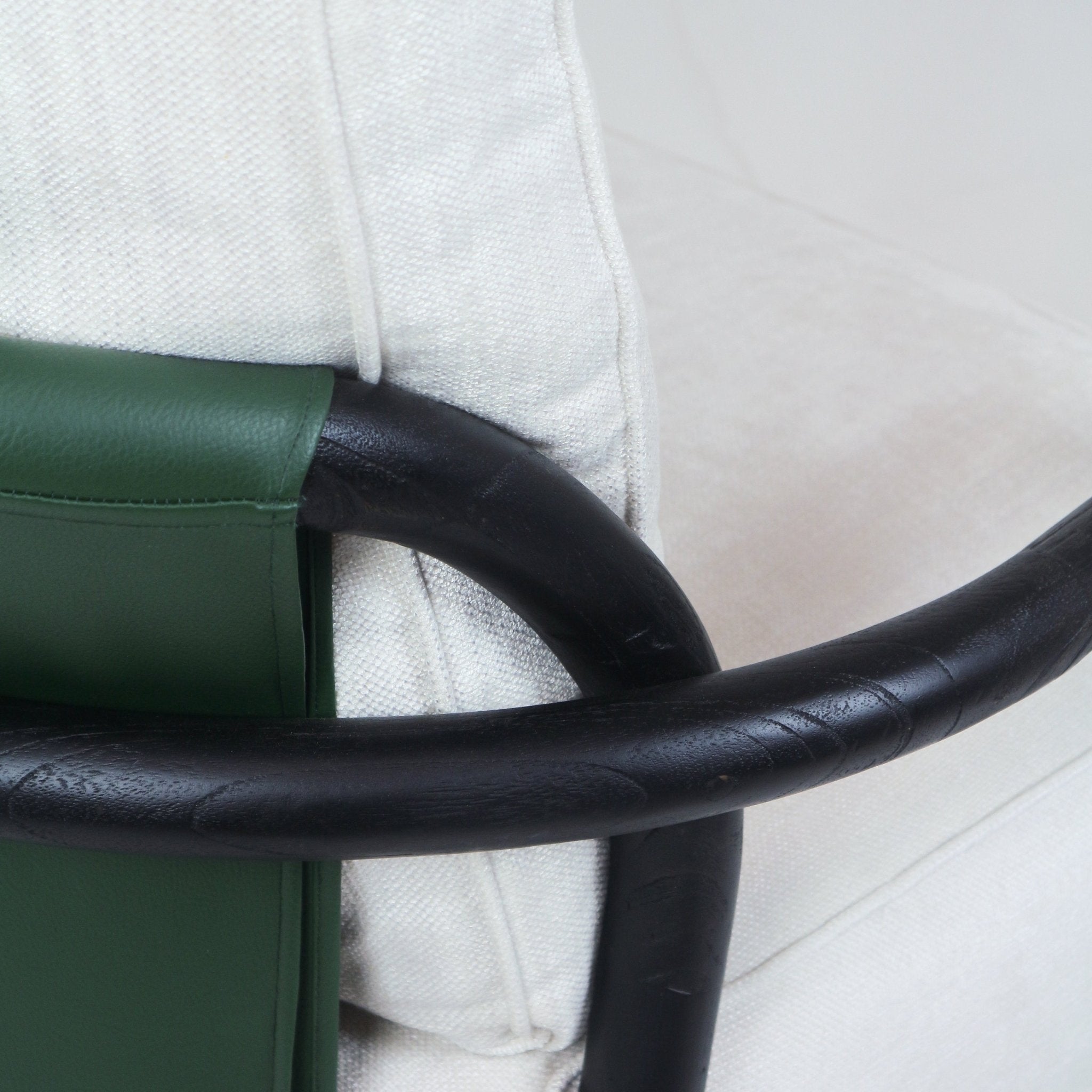 Close-up of a mid-century modern armchair showing a curved dark teak wood arm with a green leather back strap and an off-white textured fabric cushion.