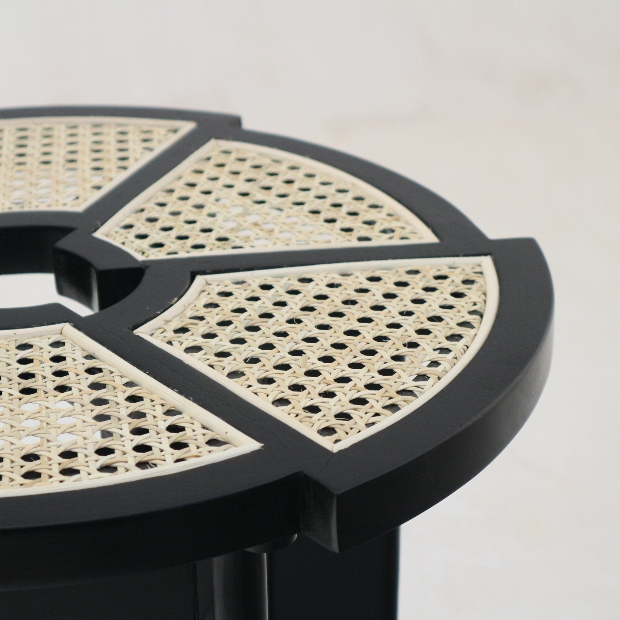 Close-up of a round side table top with natural cane rattan insets arranged in segmented panels set into a black gloss solid wood frame, showing the woven cane pattern and smooth lacquered edge.
