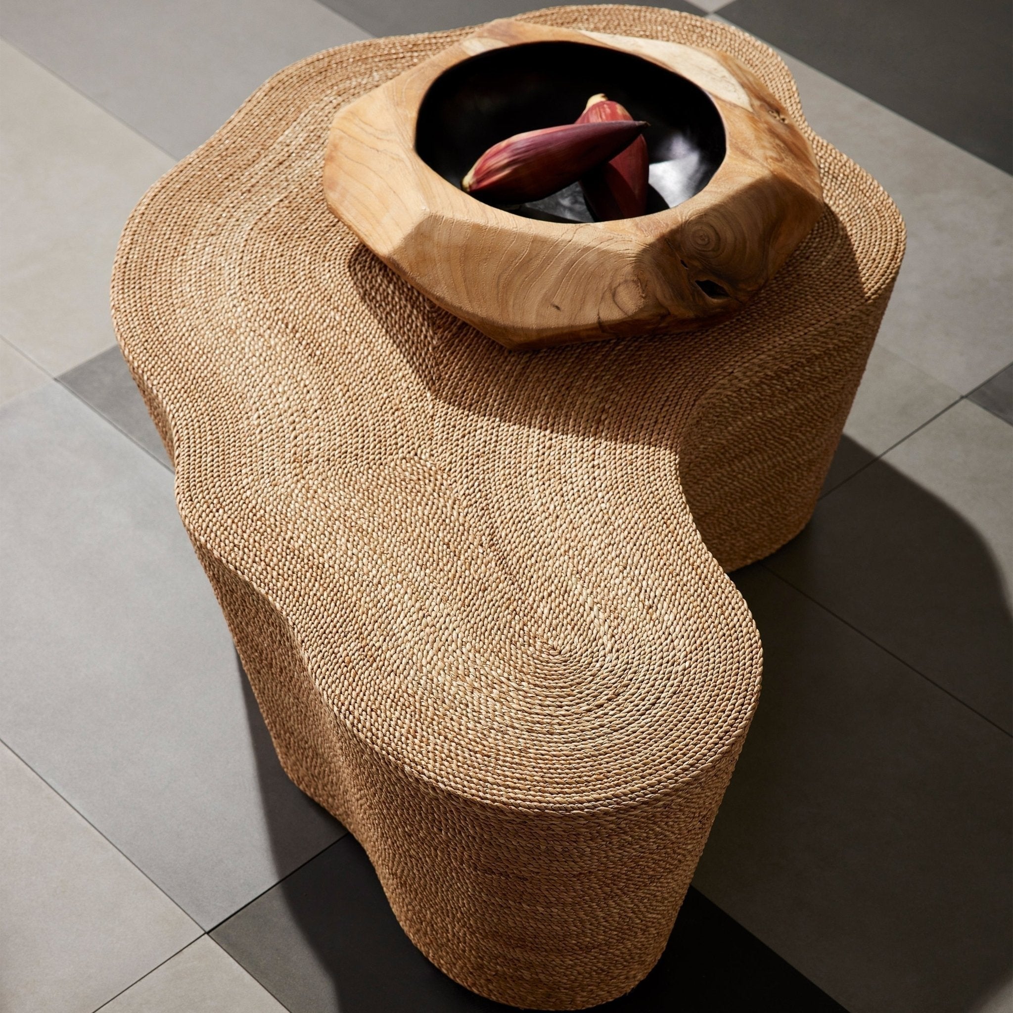 Top-down view of a natural woven abaca coffee table with an organic sculptural shape, topped by a carved natural wood bowl holding deep purple banana blossoms on a gray tiled floor.