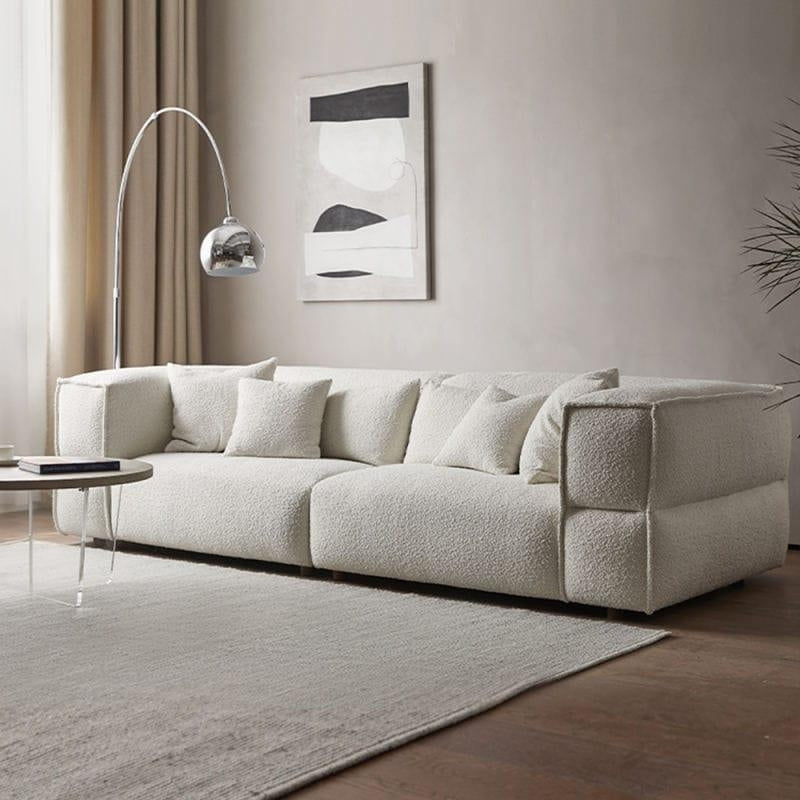 Low-profile white boucle fabric sectional sofa with plush cushions and low wooden legs in a neutral living room, styled with a chrome arc floor lamp, round coffee table, abstract wall art and textured rug.