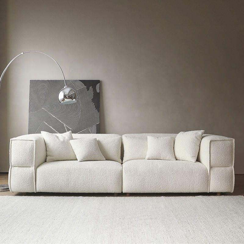 Off-white boucle sectional sofa with low wooden legs and several matching cushions, styled in a minimalist living room with a neutral wall, leaning abstract black-and-white artwork and an arched chrome floor lamp.