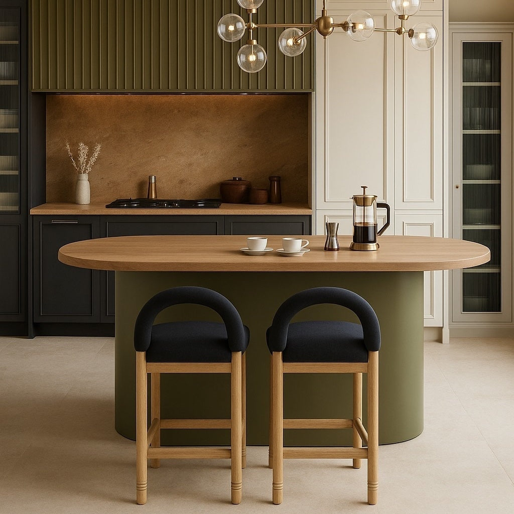 Two oak-framed counter stools with dark charcoal boucle seats and rounded low backs sit at an oval oak-topped island with an olive-green base in a contemporary kitchen, a French press and two cups on the countertop.