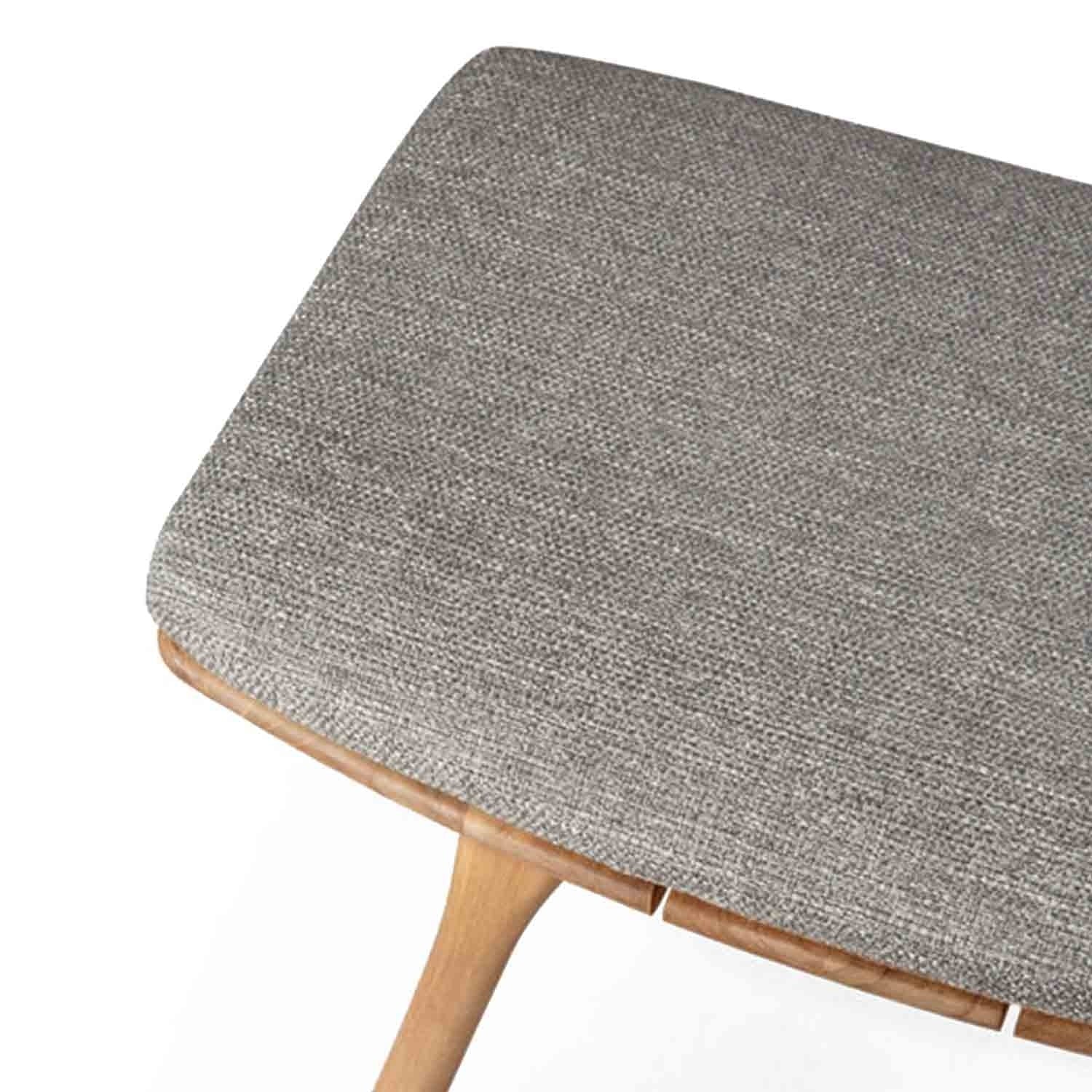 Close-up of an upholstered backless shower stool seat showing grey-beige textured fabric over a rounded teak edge and a tapered teak leg, coastal-style solid wood seating.
