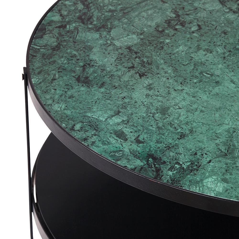 Close-up of a round green marble coffee table top with dark veining set in a thin black metal rim above a matte black lower shelf and slim metal supports.