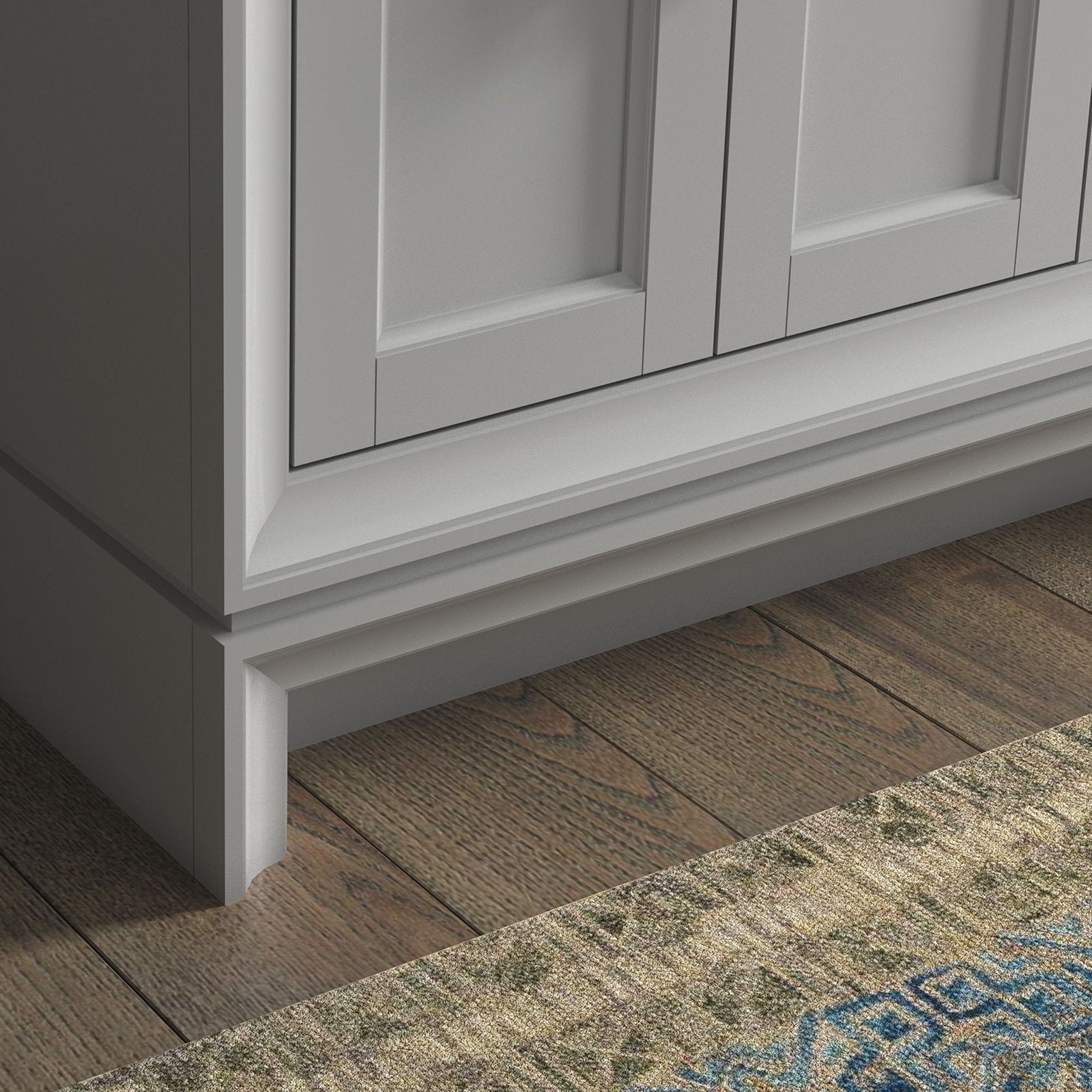 Close-up of the stone-colored solid wood bathroom vanity lower corner showing recessed panel doors, layered base molding and a toe-kick resting on hardwood floors with a patterned rug edge.
