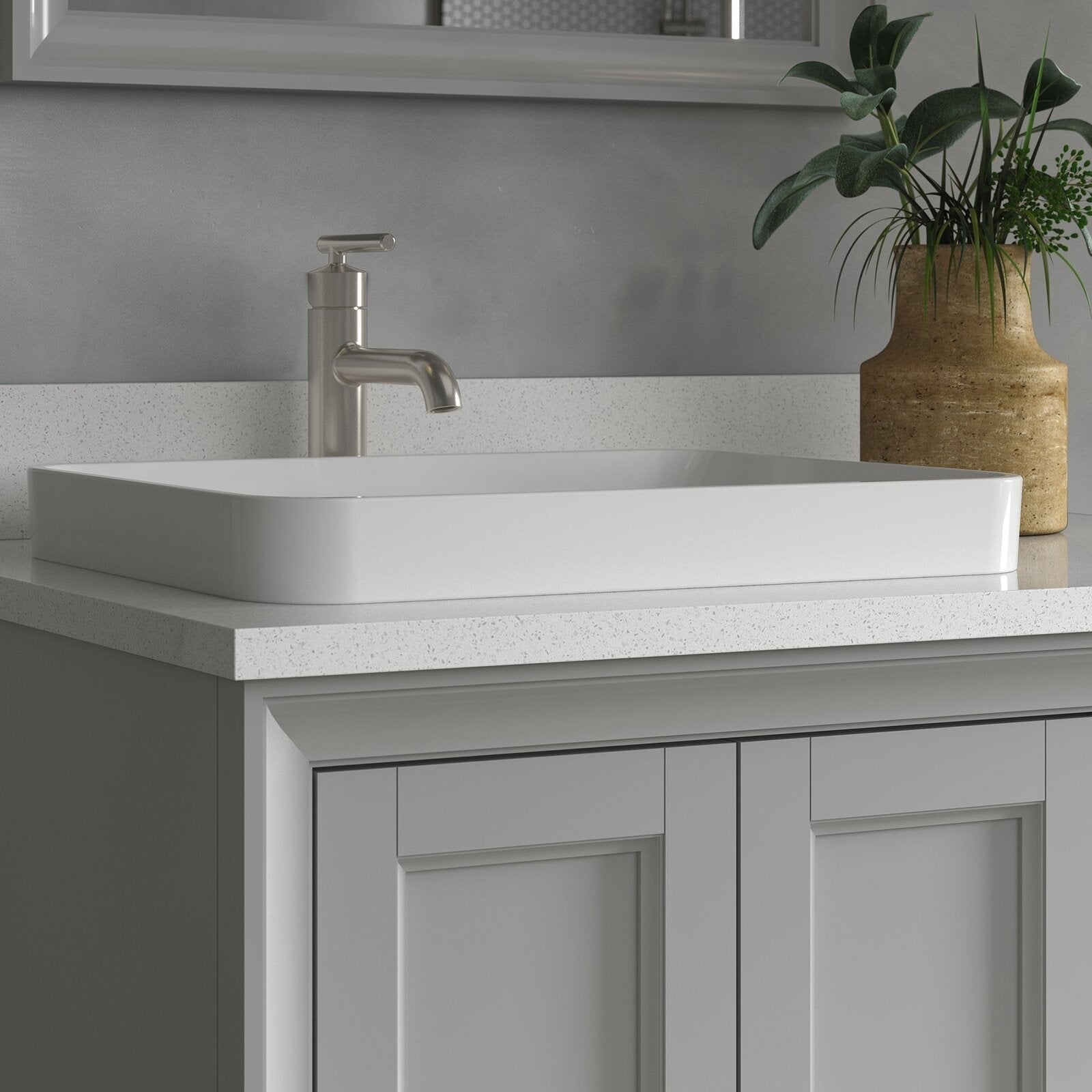 Rectangular white ceramic vessel sink sitting on a speckled stone countertop above a gray solid wood modern vanity with recessed-panel doors, paired with a brushed-nickel single-handle faucet and a brown vase with green foliage.