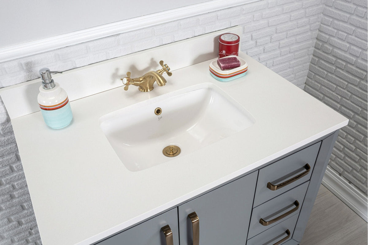 Modern gray solid-wood single-sink vanity with a white stone countertop and integrated ceramic sink, brass faucet and hardware, soap dispenser and dish on a white brick backsplash.