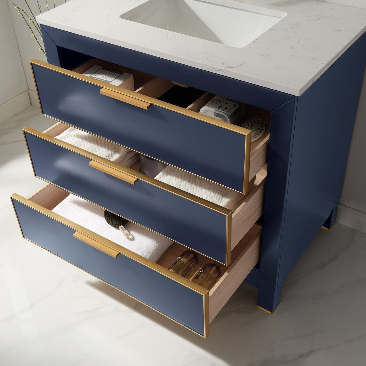 Freestanding 36-inch modern bathroom vanity with a white stone top and integrated ceramic sink, navy blue drawer fronts with gold trim and handles, three open drawers showing solid wood interiors and folded towels.