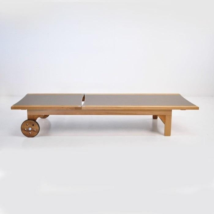 Low-profile outdoor sun lounger with a natural teak frame and taupe fabric sling top, featuring wooden wheels at one end and simple squared legs for a coastal-contemporary look.