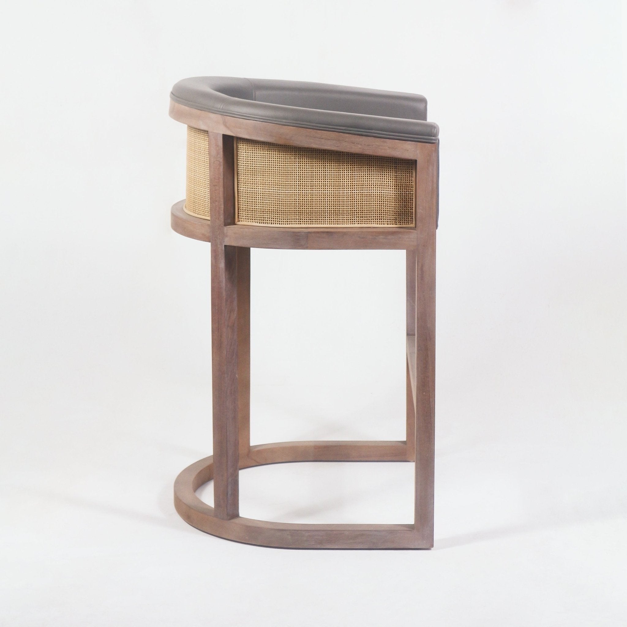 Side view of a mid-century modern teak bar stool with a curved teak frame and base, woven cane side panel, and dark faux-leather upholstered seat and low back.