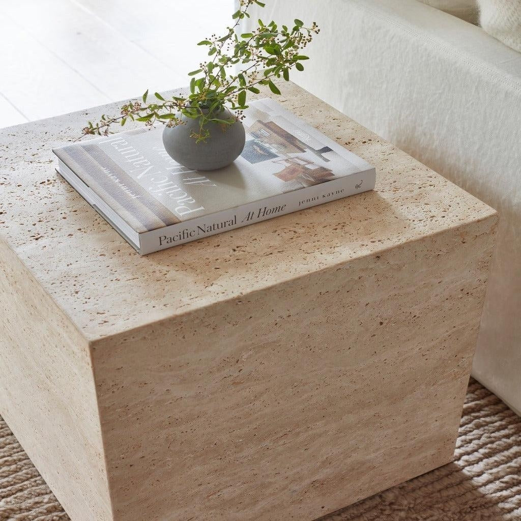 Natural beige travertine cube nightstand with visible stone texture, styled with a coffee-table book and a small gray vase holding green sprigs next to a cream sofa and textured rug.