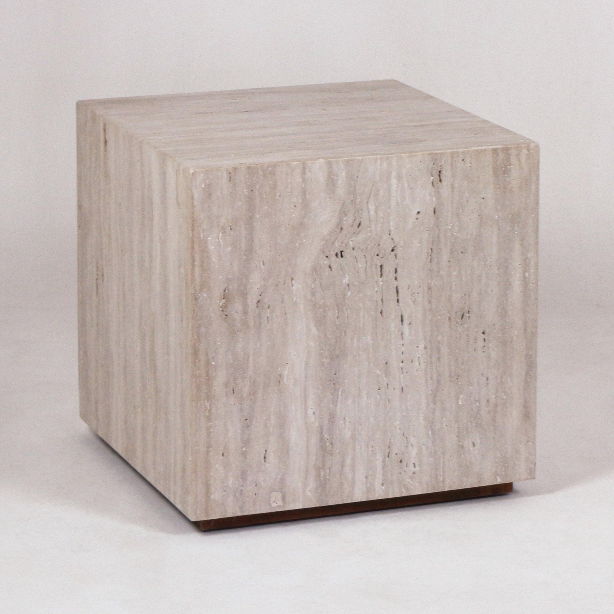 Cube-shaped travertine nightstand in a natural stone finish with subtle beige veining, shown in a three-quarter view and set on a low recessed wood plinth base.