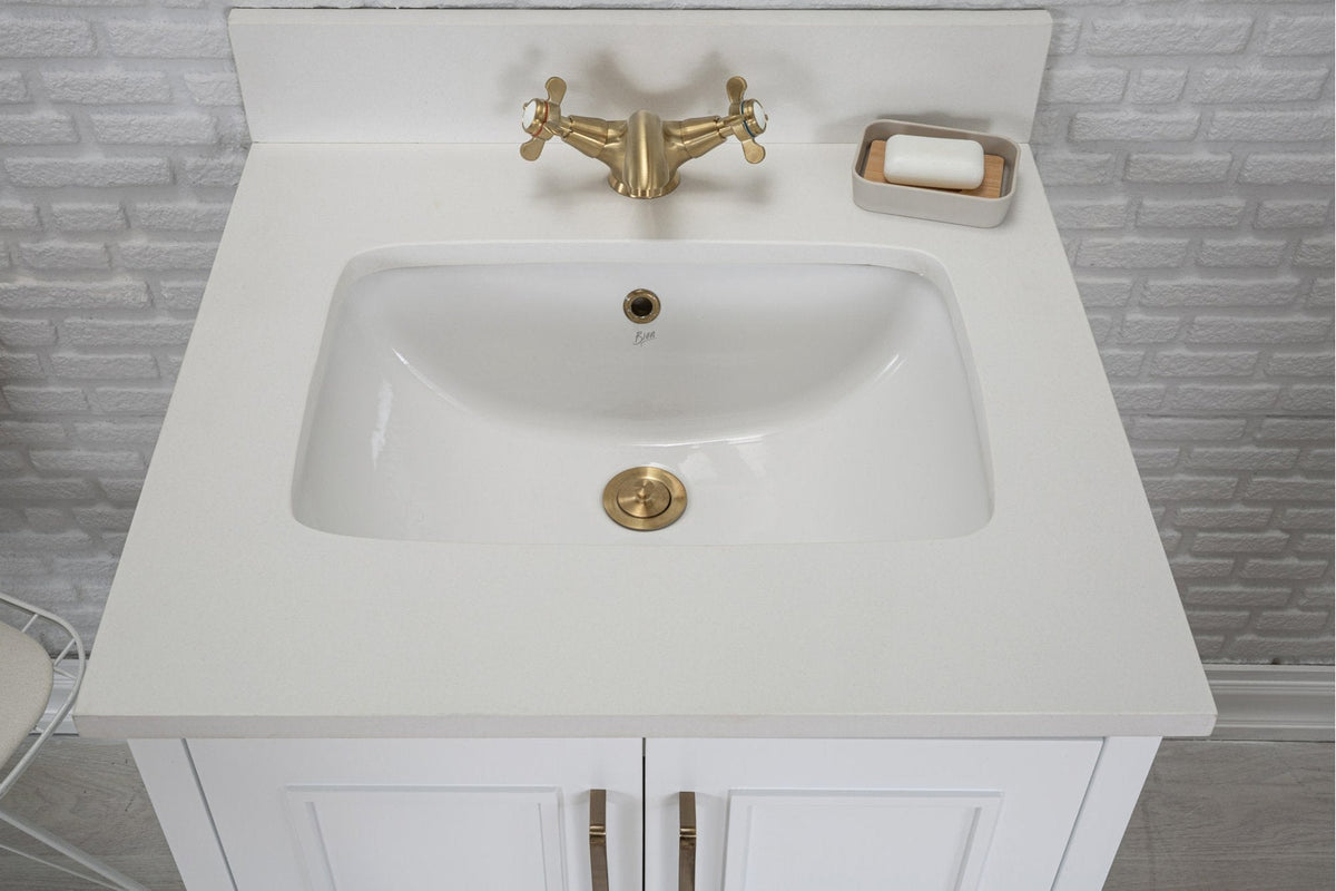 Top-down view of a modern solid wood bathroom vanity with a stone countertop, integrated white ceramic sink, brushed brass faucet and drain, soap dish at right, and white paneled cabinet doors with brass pulls.