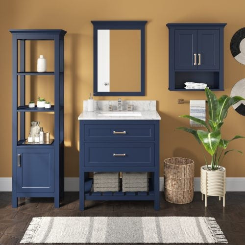 Modern navy solid wood single-sink bathroom vanity with a stone countertop and ceramic sink, shown with matching tall shelving, wall cabinet and mirror, woven laundry basket, potted plant and fringe bath rug.