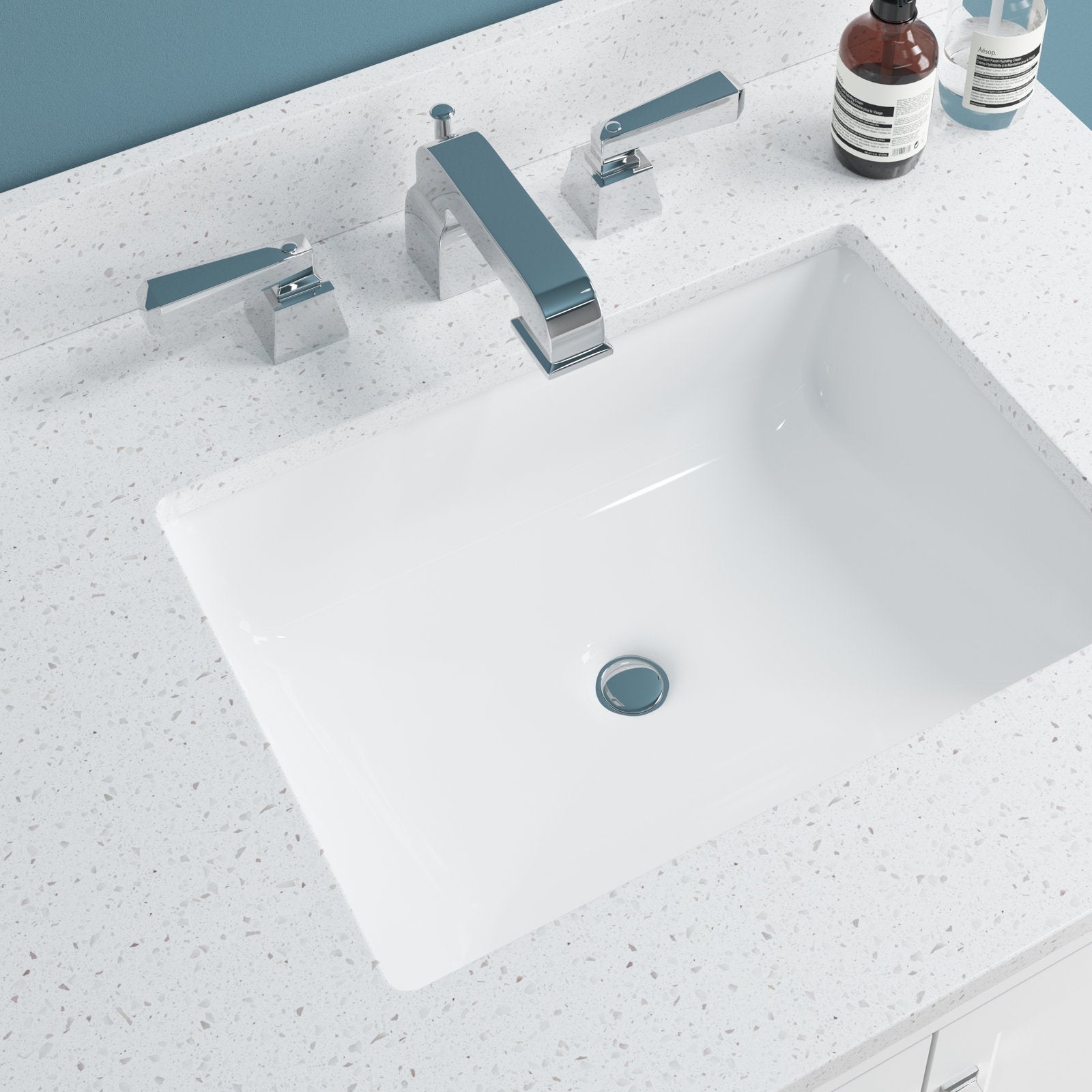 White rectangular ceramic undermount sink recessed into a speckled stone vanity top in stone color, paired with a chrome three-piece faucet and lever handles; two soap bottles rest on the back ledge against a blue wall.
