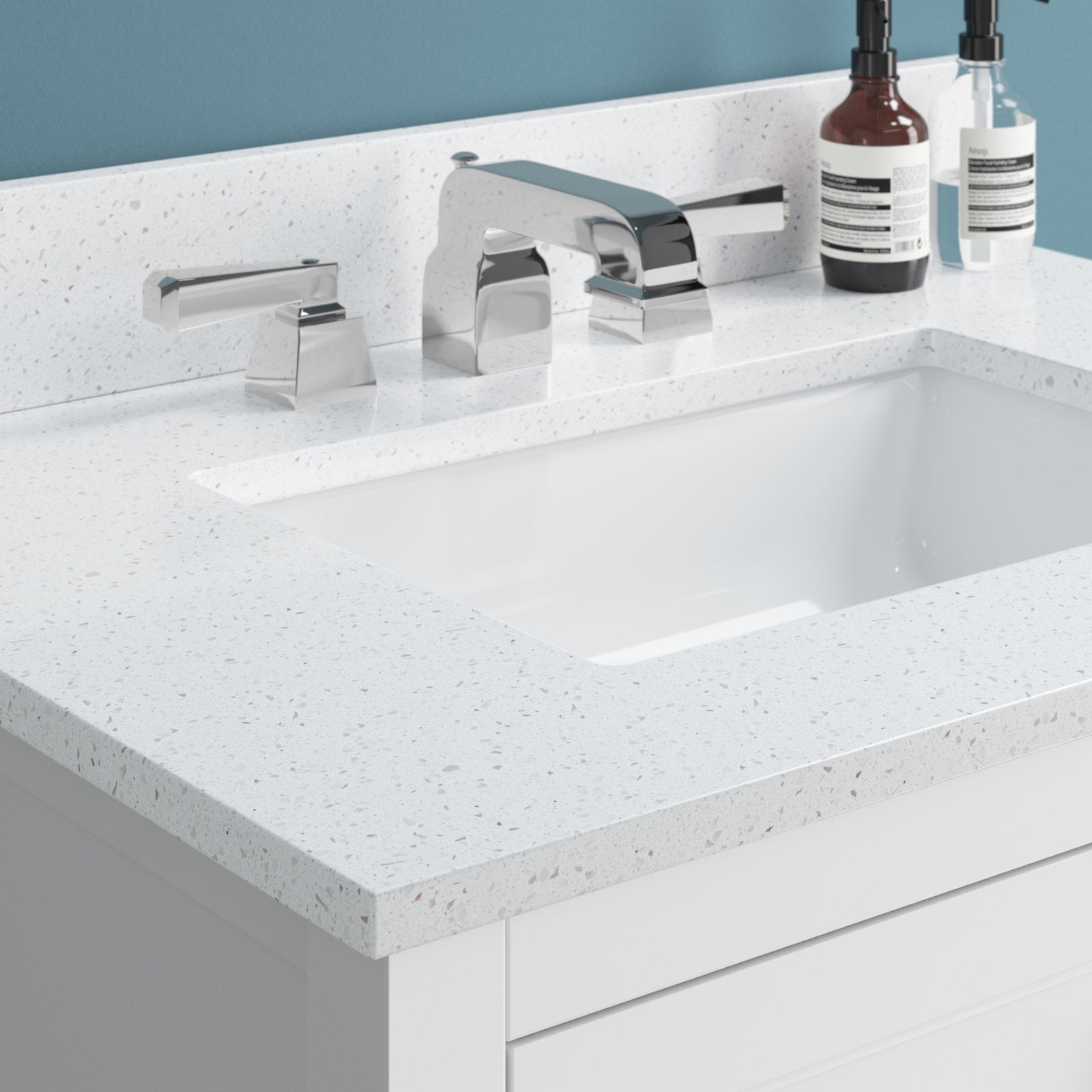Close-up of a stone-colored speckled countertop with an undermount white ceramic rectangular sink, chrome modern widespread faucet and lever handles, and white solid wood vanity drawers with soap bottles on the back