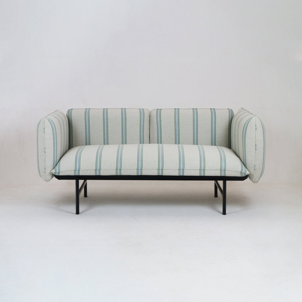 Coastal-inspired outdoor sectional sofa upholstered in light neutral striped fabric with pale green and blue vertical stripes, rounded padded arms and a low black metal frame and legs on a neutral studio background.