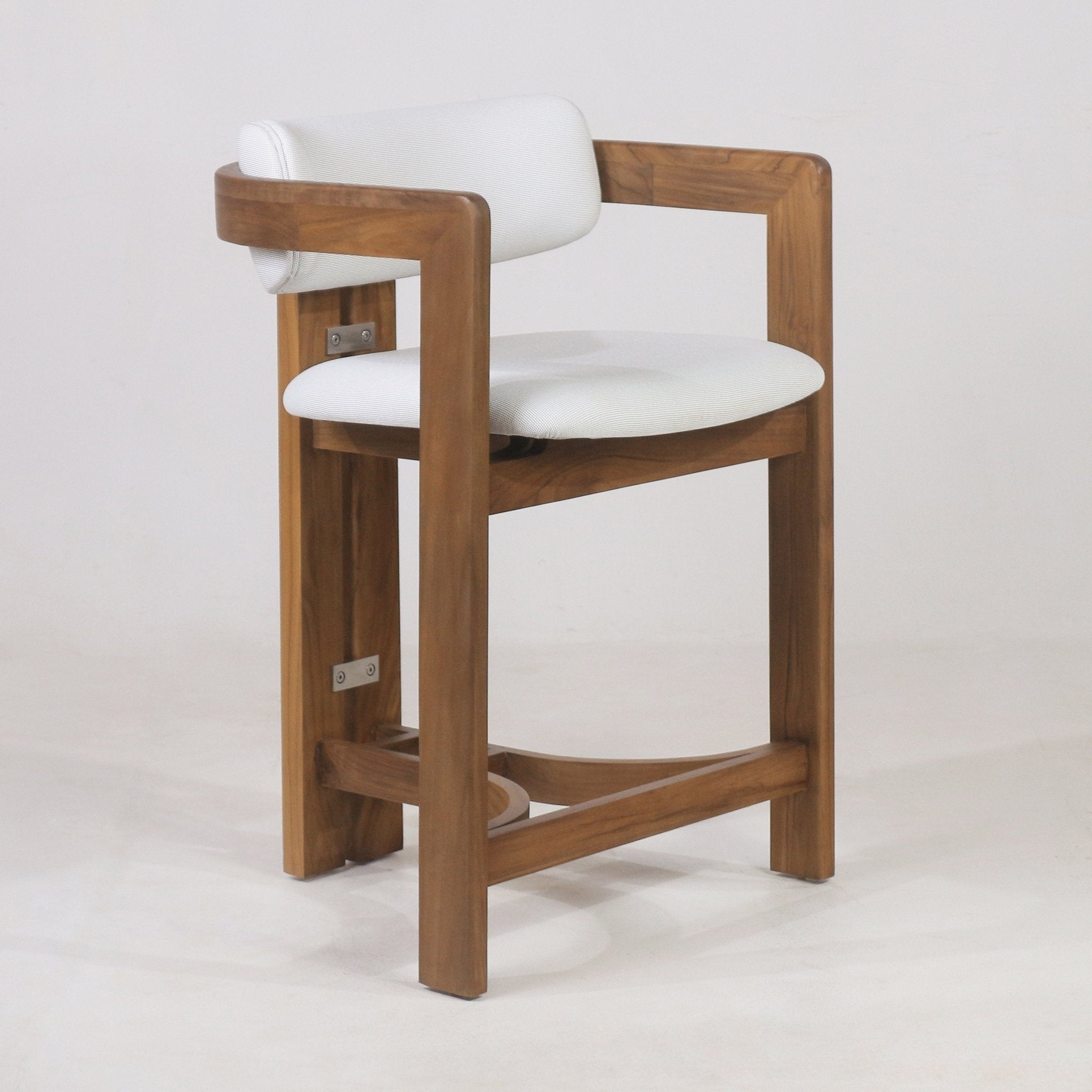 Teak outdoor bar stool with white fabric upholstered round seat and cylindrical cushioned back embraced by a curved teak arm frame, solid teak legs and integrated curved footrest in a coastal-contemporary style.