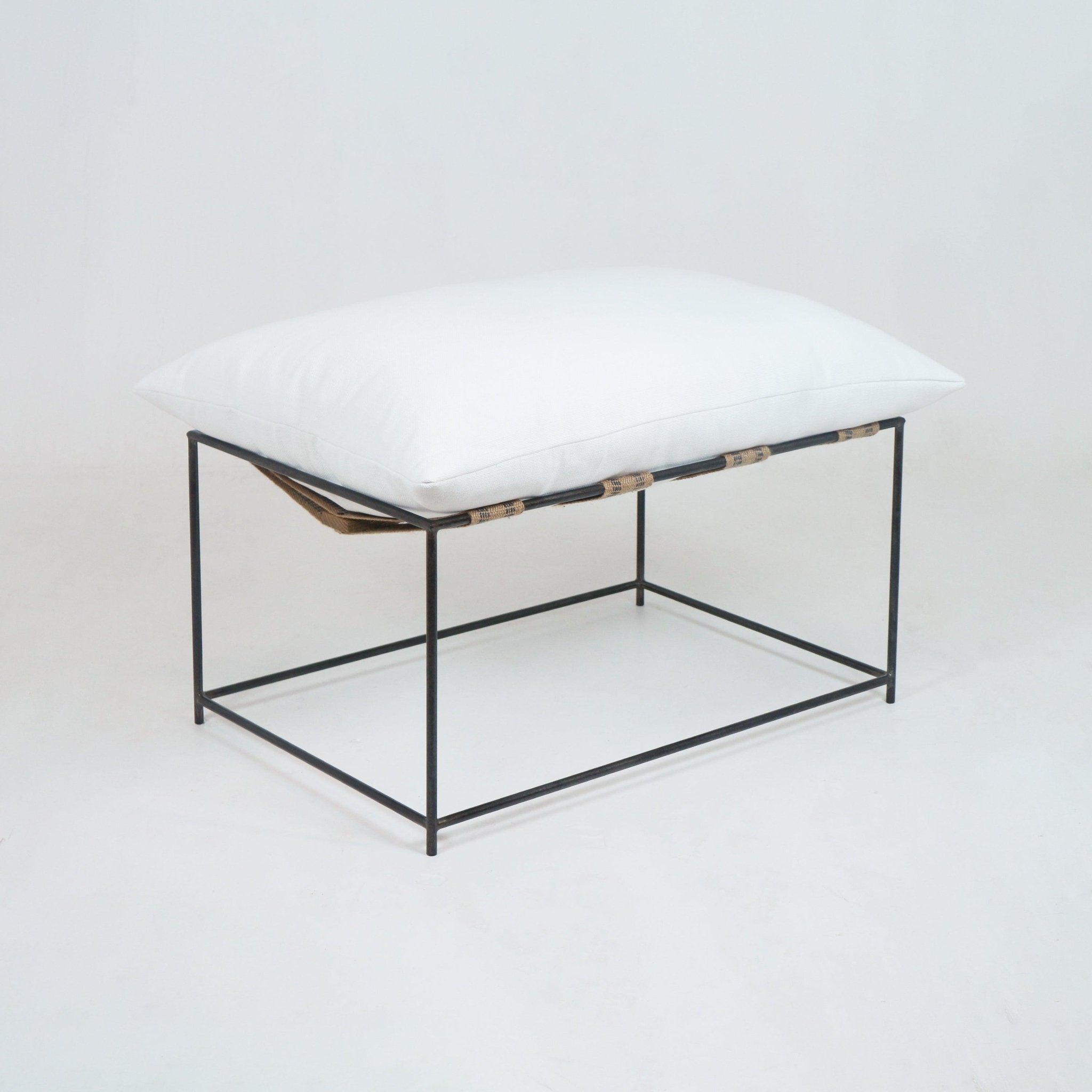 Minimalist outdoor ottoman with a plush white linen cushion seated on a slim black metal square frame and natural woven support straps, ideal for modern living or patio seating.