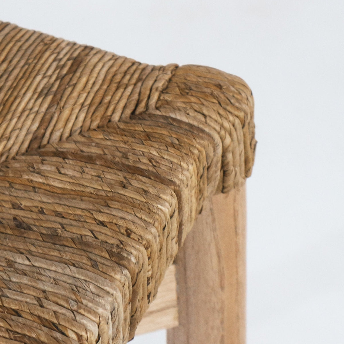 Close-up of a natural seagrass woven seat wrapped over the corner of a teak counter stool, highlighting the braided texture and warm natural teak leg.