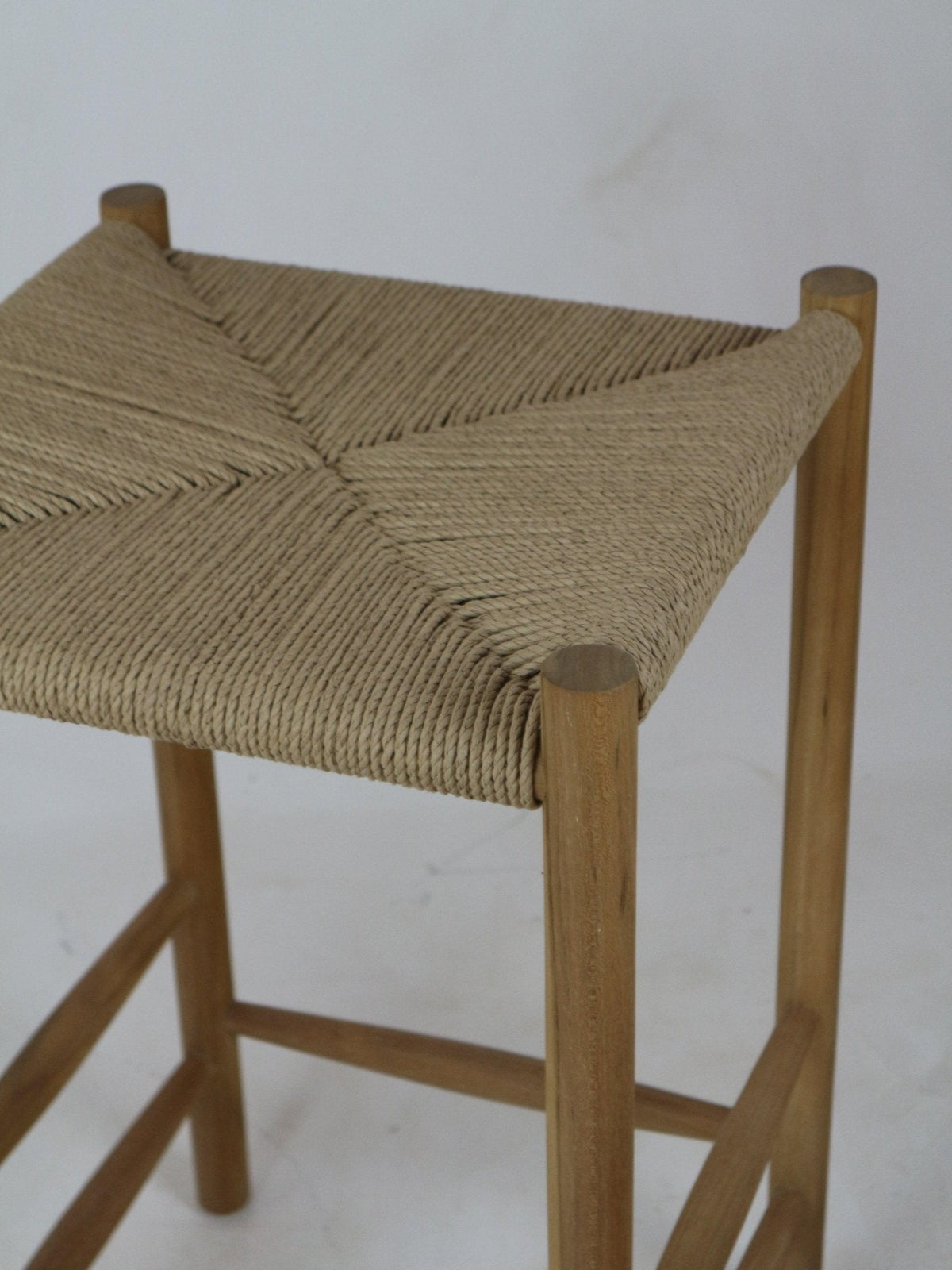 Close-up of a modern natural wood bar stool with a hand-woven beige rope seat in a diagonal square pattern, round wooden legs and integrated footrest; armless and backless on a neutral background.