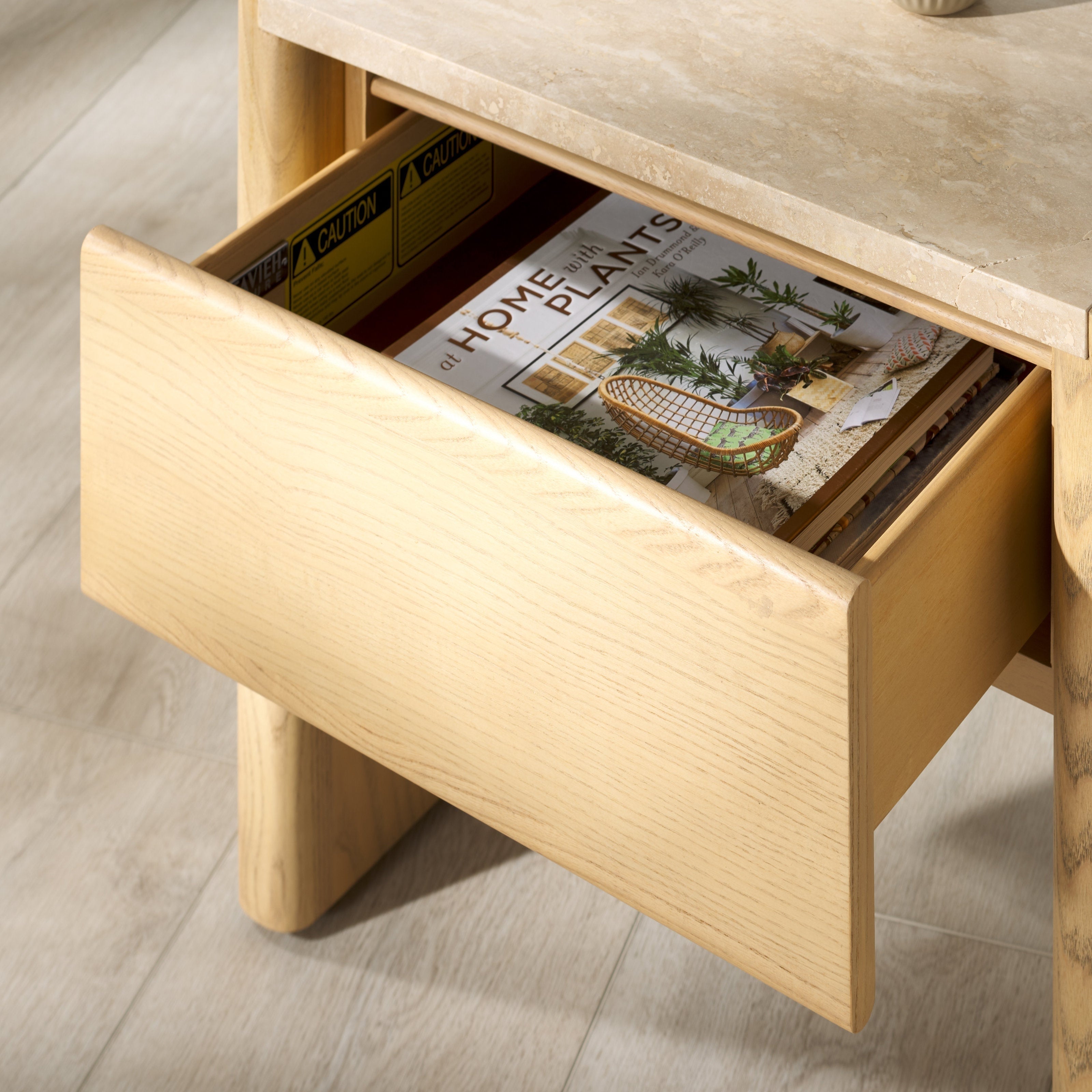 Close-up of a natural wood nightstand with an open drawer showing light oak grain, a beige marble tabletop edge above, and a lifestyle magazine tucked inside.