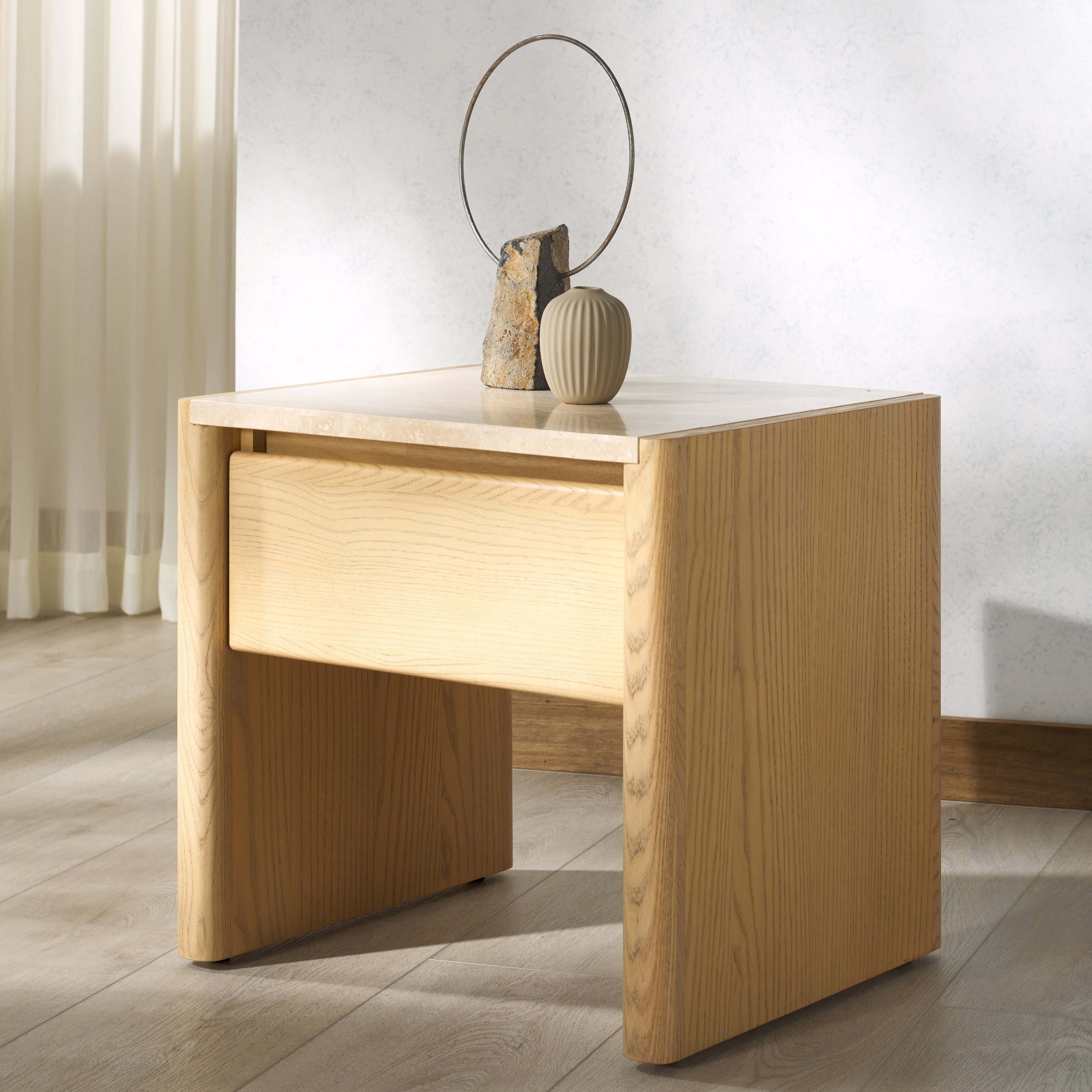 Natural wood nightstand with a light beige marble top, single recessed drawer and rounded side panels, styled with a ribbed beige vase and stone sculpture with a metal hoop in a minimalist bedroom setting.