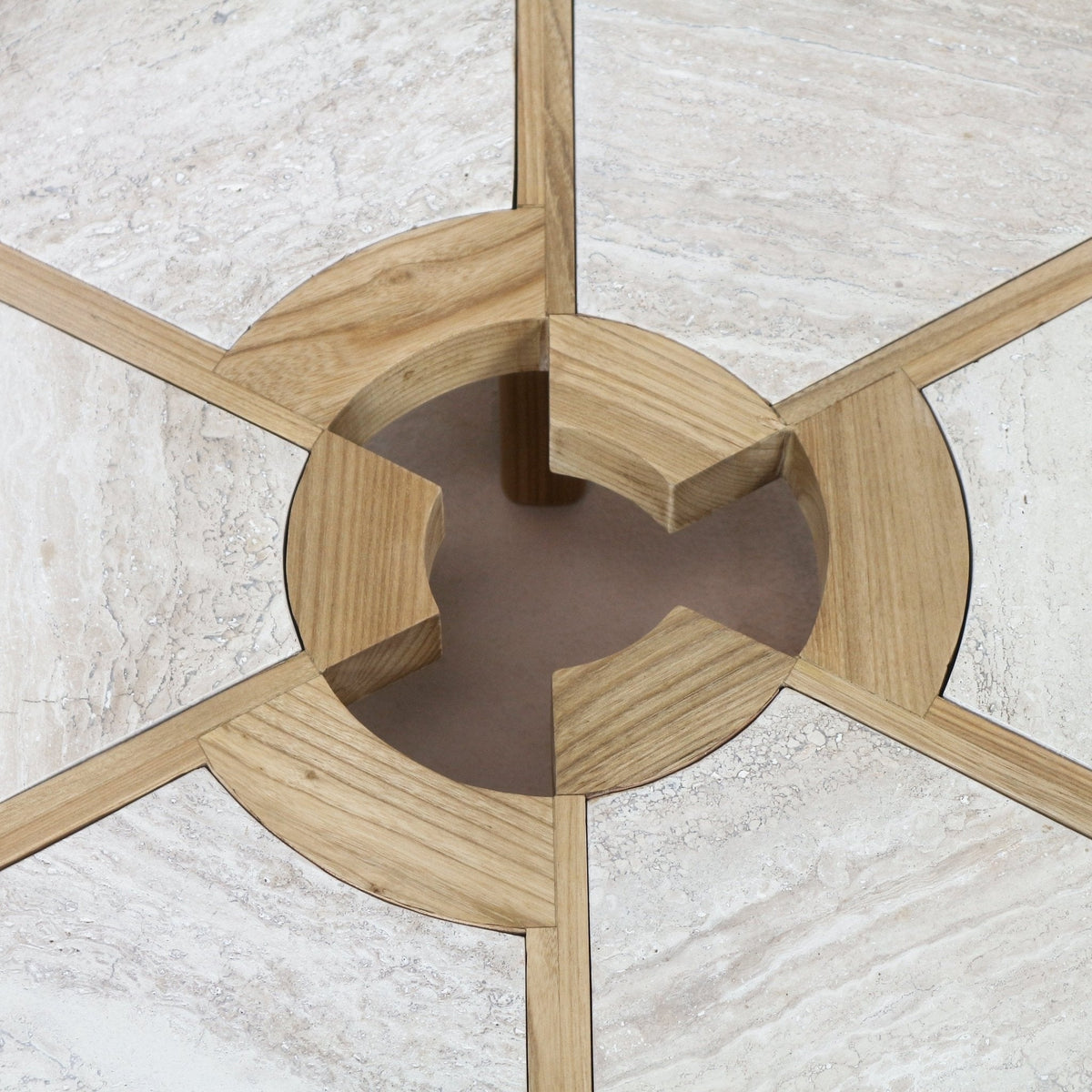 Close-up of a travertine coffee table top with natural oak inlay: light beige travertine panels divided by oak spokes forming a circular recessed center with four notched oak supports.