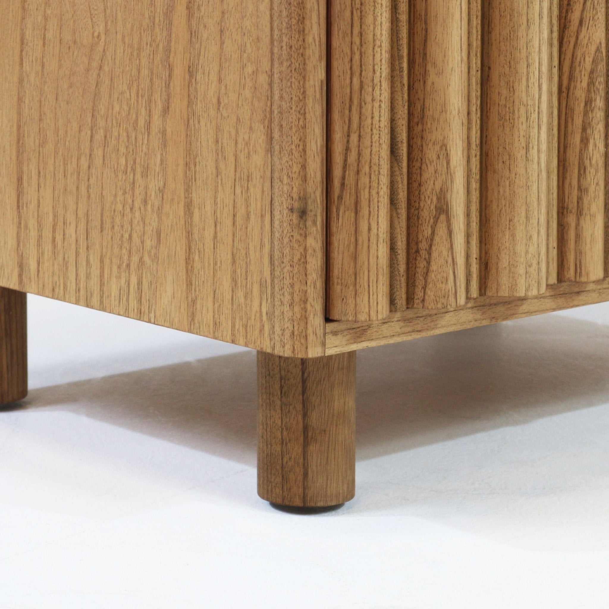 Close-up of a modern Scandinavian nightstand corner in natural solid wood, showing a fluted drawer front and round wooden leg resting on a white studio floor.