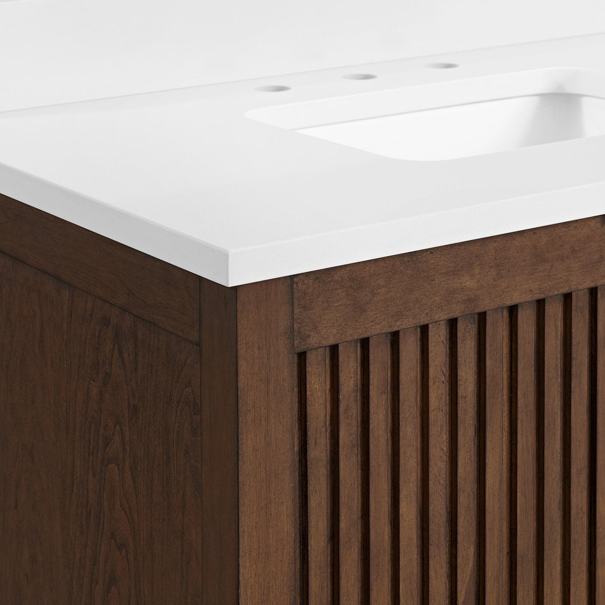 Close-up of a modern solid wood bathroom vanity corner with a white stone countertop and integrated rectangular sink, showing a fluted wooden cabinet front in warm brown natural wood finish.
