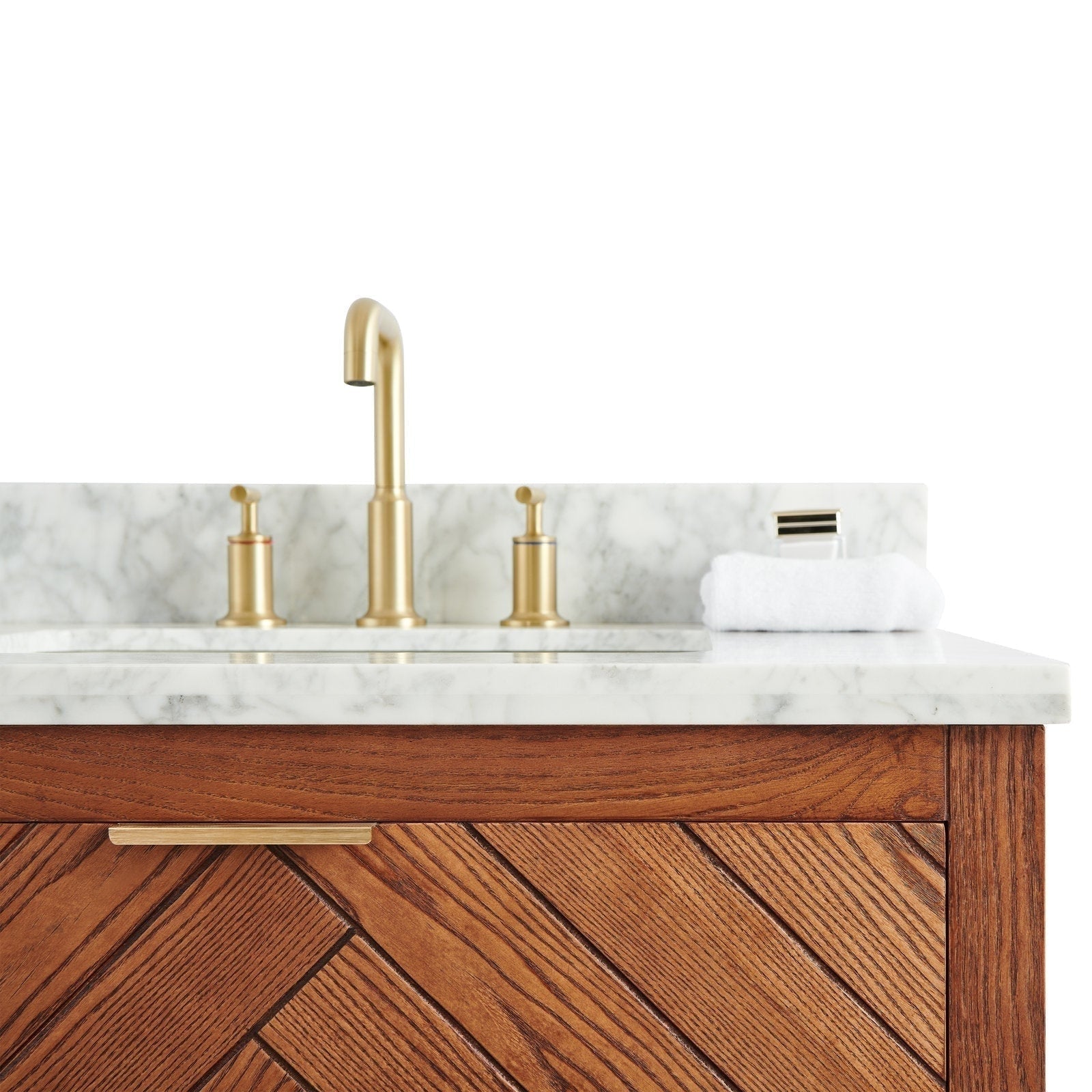 Close-up of a stone marble countertop and backsplash with brushed brass faucet and matching disc handles, a folded white towel, and a warm solid wood vanity front with diagonal chevron grain and slim brass pull.