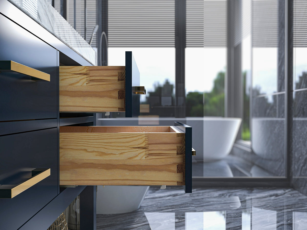 Close-up of a modern bathroom vanity showing two open solid wood dovetail drawers with dark stone-colored fronts and brushed gold handles under a stone countertop, freestanding tub and marble floor visible in background.